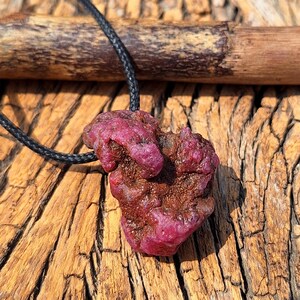 Puede incluir: Un colgante de rubí en bruto y sin pulir con una superficie texturizada de color magenta intenso y marrón rojizo. La piedra preciosa de forma orgánica irregular está suspendida de un cordón trenzado negro fino, mostrando formaciones minerales.