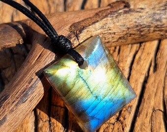 Puede incluir: Colgante cuadrado de labradorita con cordón negro. La piedra preciosa muestra tonos iridiscentes azules, verdes y amarillos. El colgante se exhibe sobre una superficie de madera.