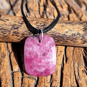Peut inclure: Pendentif rectangulaire en pierre rose sur un collier en cordon noir. Le pendentif présente un aspect marbré. Le collier est présenté sur un morceau de bois avec un grain naturel texturé. La pierre est d'un rose vif.