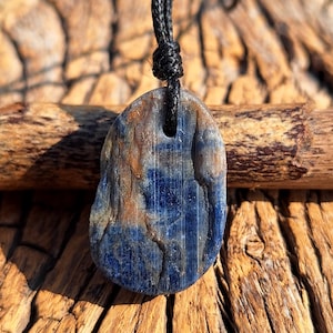 May include: A blue and brown stone pendant with a black cord. The stone has a natural, organic shape and texture, with streaks of blue and brown. The cord is knotted at the top, creating a loop for wearing.