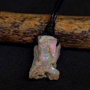 Könnte beinhalten: Ein Anhänger aus rohem Opal an einer schwarzen Kordel. Der Opal hat eine raue, natürliche Textur mit roten und blauen Lichtreflexen im Inneren. Der Anhänger hängt an einer geknoteten schwarzen Kordel, die die natürliche Schönheit des Edelsteins hervorhebt.
