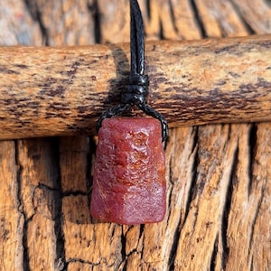 以下が含まれることがあります： 黒い革紐に吊るされた、赤褐色の原石ルビーのペンダント。宝石は、表面にテクスチャ加工が施された、ほぼ長方形の形をしています。背景は、自然で素朴な外観の木製の表面です。