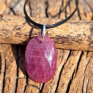 May include: A pink gemstone pendant on a black cord necklace. The pendant is faceted and has a shiny, polished finish.
