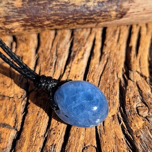 May include: A blue oval-shaped gemstone pendant on a black cord necklace. The gemstone has a translucent appearance. The necklace is displayed on a textured, brown wooden surface, with a piece of wood in the background.