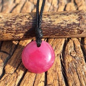 May include: A circular, polished, magenta pendant necklace with a black cord. The pendant is set against a backdrop of textured wood and a wooden log.