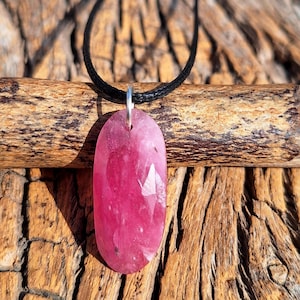 May include: A faceted, oval-shaped pink gemstone pendant suspended from a black cord necklace. The pendant is a vibrant shade of pink with visible internal inclusions. The necklace is displayed against a textured wooden background.