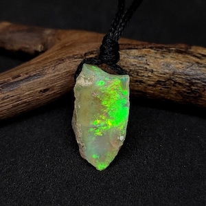 Könnte beinhalten: Ein Anhänger aus rohem, natürlichem Opal. Der Opal hat eine hellgrüne Farbe mit leuchtend grünen und gelben Lichtreflexen. Der Anhänger hängt an einer schwarzen Kordel.