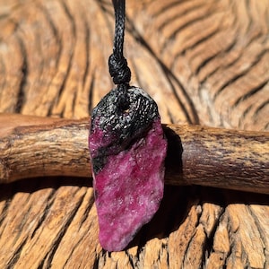 May include: A raw, irregularly shaped pendant featuring a vibrant magenta-pink stone section transitioning into a dark, textured black top. The pendant is suspended from a thin, black braided cord that is secured with a simple knot at the top.