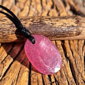 May include: A pink oval pendant necklace with a black cord. The pendant is a translucent pink stone with a faceted edge. The necklace is displayed on a wooden surface.