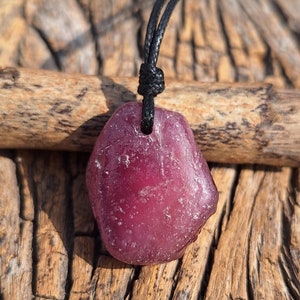 May include: A deep red, irregularly shaped pendant necklace. The pendant is suspended from a black cord, knotted at the top. The background is a textured, weathered wood surface.