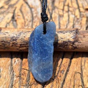 May include: A blue stone pendant with a black cord. The stone has a rough, irregular shape and a deep blue colour with lighter areas. The pendant is suspended against a wooden background.