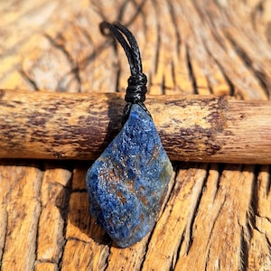 Può includere: Un ciondolo di pietra preziosa blu grezza con cordino nero. La pietra dalla forma irregolare ha una consistenza ruvida e mostra sfumature di blu con accenni di marrone. Il ciondolo è sospeso da un cordino nero annodato, creando un'estetica naturale.