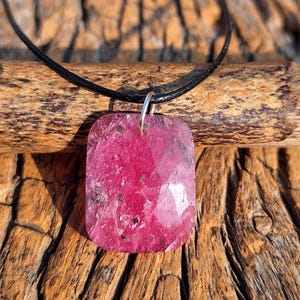 May include: A faceted, rectangular pendant necklace featuring a vibrant pink gemstone. The gemstone is suspended from a black cord with a silver-colored bail. The necklace is displayed on a wooden surface.