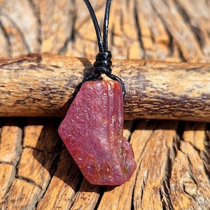 以下が含まれることがあります： 黒い紐に吊るされた、生の天然ルビーのペンダント。ルビーは深い赤色で、目に見える質感とバリエーションがあります。ペンダントは紐に結び付けられており、シンプルで素朴なネックレスになっています。背景は木製の表面です。