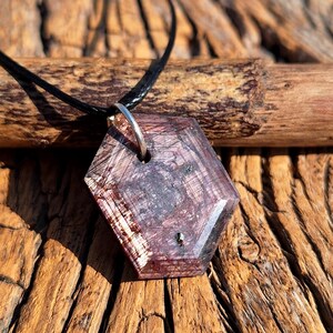 May include: A hexagonal pendant necklace with a dark purple and reddish-brown stone. The stone has a unique pattern and is attached to a black cord with a silver-colored ring. The necklace is displayed on a wooden surface.