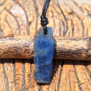 Può includere: Un pendente rettangolare grezzo di kianite blu, sospeso a un cordino di cuoio nero. La kianite mostra una texture naturale a strati con varie tonalità di blu. Il pendente è posto su uno sfondo di legno naturale.
