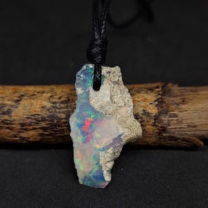 Könnte beinhalten: Ein roher Opal-Anhänger mit einer schwarzen Kordelkette. Der Opal ist hellgrau mit blauen, grünen und roten Schimmern.
