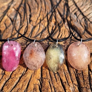 May include: Four gemstone pendants on black cords. The pendants are in shades of pink, peach, and yellow, with a variety of shapes. The background is a textured wooden surface.
