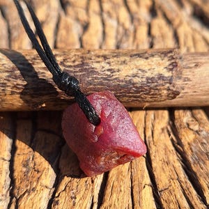 May include: A raw, deep red gemstone pendant suspended from a black cord. The irregularly shaped stone has a rough, textured surface. The cord is knotted at the top, creating a necklace. The background is a wooden surface.