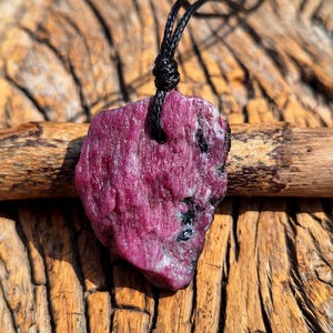 May include: A rough-cut ruby pendant on a black cord. The ruby is a deep pink-red hue, featuring black inclusions and a textured surface. The pendant hangs from a knotted black cord, highlighting the natural characteristics of the gemstone.