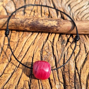 Puede incluir: Un collar hecho a mano con un colgante de piedra preciosa roja pulida. El colgante tiene forma cuadrada redondeada, suspendido de un collar de cordón negro. El cordón está anudado a cada lado del colgante. El collar descansa sobre una superficie de madera texturizada.