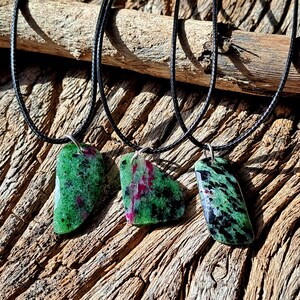 May include: Three green, black, and pink gemstone pendants on black cords. The pendants are all different shapes and sizes. The cords are hanging from a piece of driftwood.