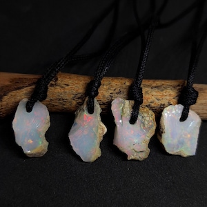 Può includere: Cinque ciondoli in opale iridescente su cordini neri, ognuno con una forma e un motivo unici. I ciondoli sono esposti su un pezzo di legno galleggiante.