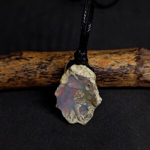 Könnte beinhalten: Ein roher Opal-Anhänger mit einer schwarzen Schnurkette. Der Opal ist milchig weiß mit Schimmern in Rot, Grün und Blau.