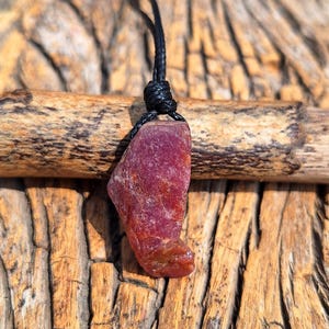 May include: A raw, uncut ruby pendant suspended from a black cord. The ruby is a deep red color with a rough, textured surface. The pendant is displayed against a wooden background, highlighting the natural beauty of the gemstone.