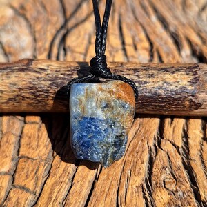 Può includere: Un ciondolo in pietra naturale con un cordino nero. La pietra presenta una miscela di colori blu, marrone e bianco. Il ciondolo è sospeso a un cordino nero annodato, mettendo in risalto la consistenza e le variazioni di colore uniche della pietra.