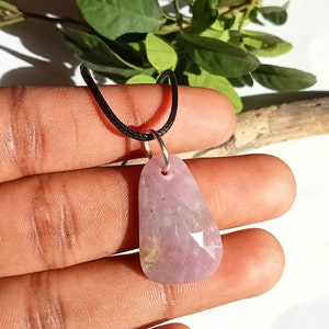 May include: A light pink, faceted pendant on a black cord necklace. The pendant is teardrop-shaped with a silver-coloured bail. The stone has a translucent quality and is suspended by a black cord.
