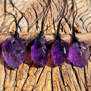 May include: Four raw amethyst crystal pendants with black cords. The purple crystals are rough-cut and vary in size and shape. The pendants are displayed on a wooden surface with a natural, rustic appearance.