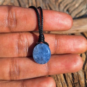 May include: A blue oval gemstone pendant on a black cord. The gemstone has a cloudy appearance and is suspended from a knotted cord. The pendant is held up by a person's fingers.