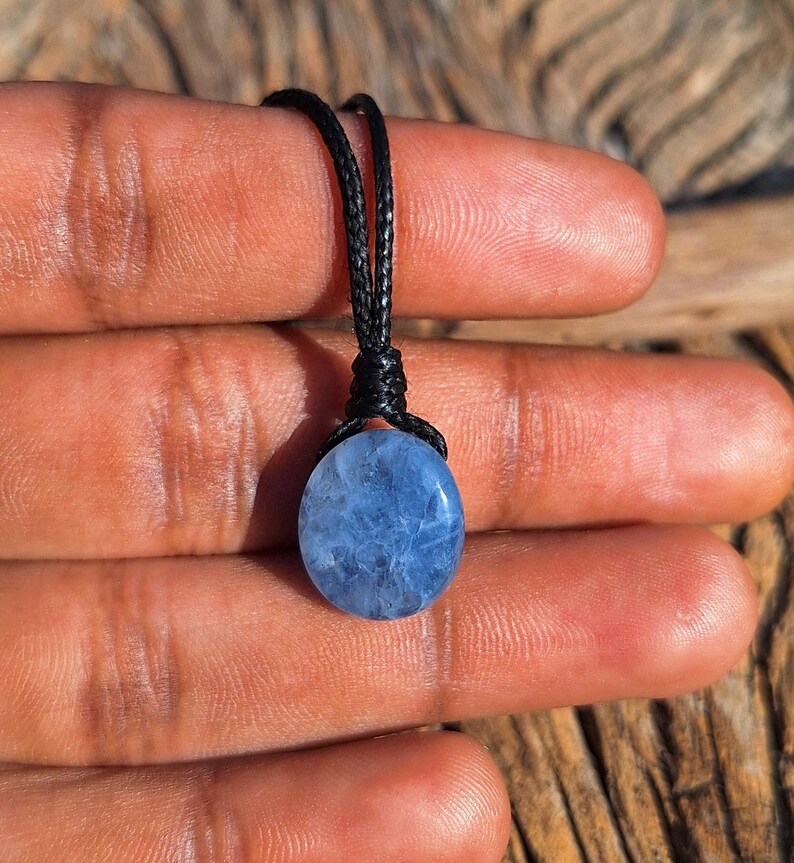 May include: A blue oval gemstone pendant on a black cord. The gemstone has a cloudy appearance and is suspended from a knotted cord. The pendant is held up by a person's fingers.