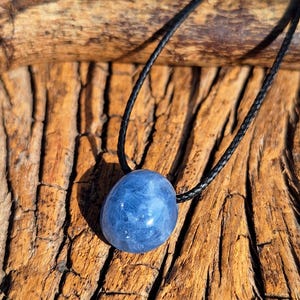 May include: A blue gemstone pendant on a black cord. The polished, oval-shaped stone has a cloudy appearance. The cord is knotted at the top and is set against a textured, brown wooden surface. The pendant is a natural, organic accessory.