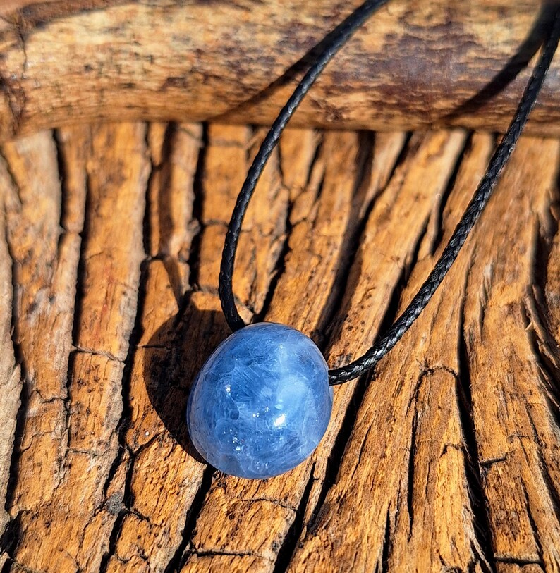 May include: A blue gemstone pendant on a black cord. The polished, oval-shaped stone has a cloudy appearance. The cord is knotted at the top and is set against a textured, brown wooden surface. The pendant is a natural, organic accessory.