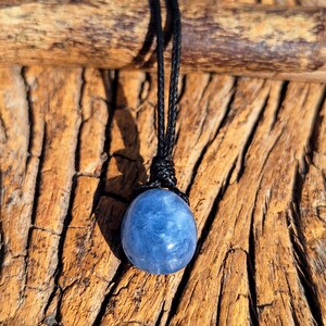 May include: A blue sphere pendant necklace with a black cord. The gemstone is a light blue color and appears to be polished. The necklace is displayed against a textured, brown wooden background.