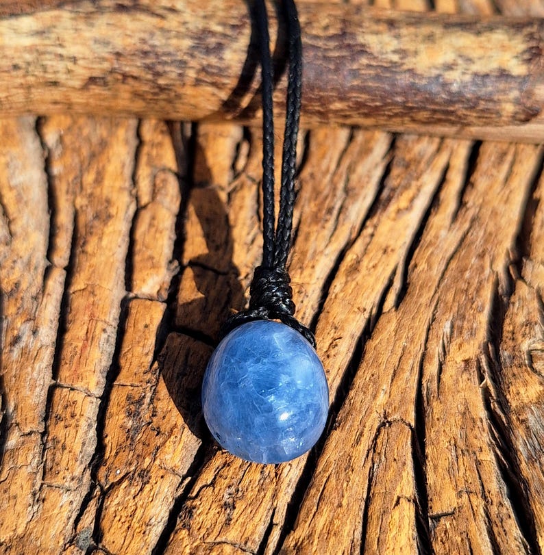 May include: A blue sphere pendant necklace with a black cord. The gemstone is a light blue color and appears to be polished. The necklace is displayed against a textured, brown wooden background.