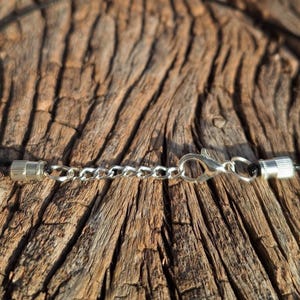 May include: Close-up of a necklace clasp and chain. The necklace features a black cord with silver-colored metal clasps and a chain for adjusting the length. The background is a textured, brown wooden surface.