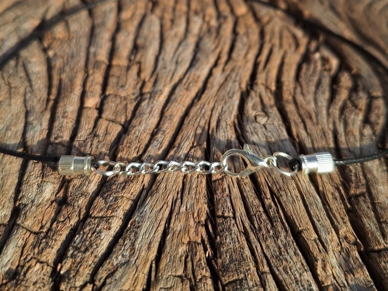 May include: Close-up of a necklace clasp and chain. The necklace features a black cord with silver-colored metal clasps and a chain for adjusting the length. The background is a textured, brown wooden surface.