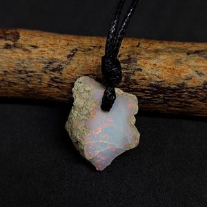 Könnte beinhalten: Ein roher, weißer Opal-Anhänger mit einer rauen, unregelmäßigen Form und einer schwarzen Schnur. Der Opal hat einen leichten rosa Farbton und ein schimmerndes, irisierendes Finish.