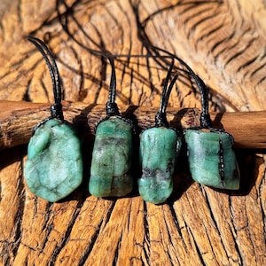 May include: Four raw emerald pendants, each featuring an irregular, unpolished green stone with natural dark inclusions. Every pendant is securely attached to a thin, black cord by a simple knot, showcasing the organic texture of the gemstones.