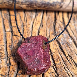 May include: A raw, uncut ruby gemstone pendant. The deep red stone is suspended from a black cord, showcasing its natural, textured surface. The pendant is displayed against a rustic wooden background, highlighting the stone's color and form.