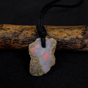 Könnte beinhalten: Ein roher Opal-Anhänger mit schwarzem Band. Der Opal hat eine unregelmäßige, natürliche Form mit weißen, roten und grünen Farbtönen. Der Anhänger hängt an einem schwarzen, geflochtenen Band und bildet eine einzigartige Halskette.