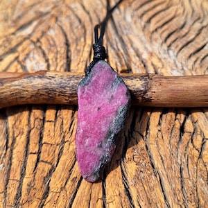 May include: A raw, unpolished pink and black gemstone pendant suspended from a thin black cord. The stone features a vibrant, textured magenta center with dark, rough edges, showcasing a natural, irregular shape and crystalline surface structure.