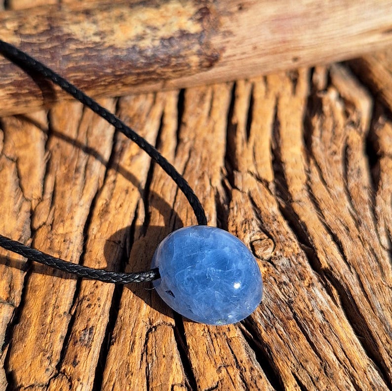 May include: A blue oval-shaped pendant necklace. The pendant is a light blue color and appears to be a polished stone. The necklace cord is black and braided. The background is a textured, brown wooden surface.