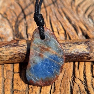 May include: A blue and brown stone pendant, suspended from a black cord. The pendant has a polished surface and an irregular shape. The necklace is displayed against a wooden background. The stone is a natural product.