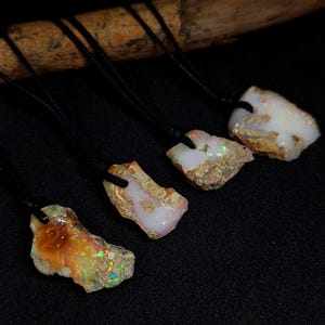 Könnte beinhalten: Fünf Opal-Anhänger an schwarzen Schnüren. Die Opale sind roh und haben eine weiße, milchige und irisierende Optik. Die Anhänger sind in einer Reihe auf einer dunklen Oberfläche angeordnet.