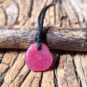Könnte beinhalten: Ein Anhänger mit einem glatten, durchscheinenden, leuchtend rosa Stein in unregelmäßiger, abgerundeter Form. Der Stein hängt an einer dünnen, schwarzen Kordel, die durch ein Loch an der Oberseite geführt und mit einem Knoten fixiert ist.