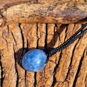 May include: A blue oval-shaped pendant necklace with a black cord. The pendant has a textured surface and is suspended from a braided black cord. The necklace is displayed on a wooden surface.